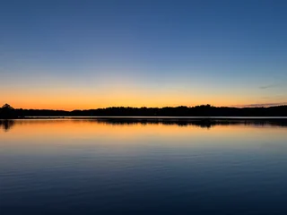 Orange sunset reflected on a lake beneath a blue sky and a dark, tree-lined shore.